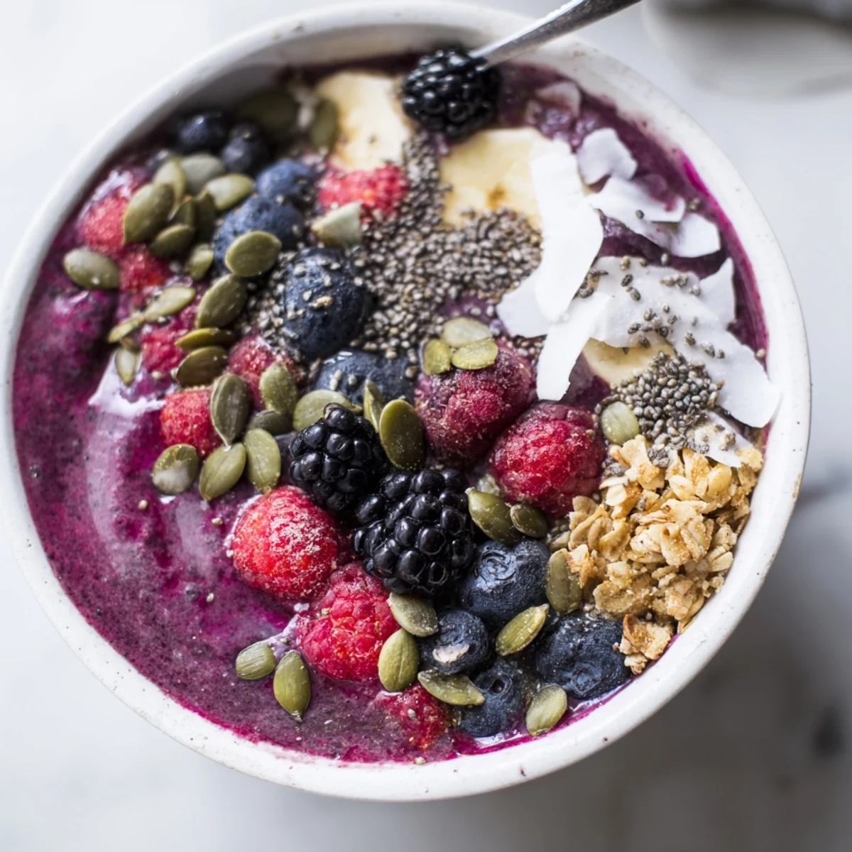 The thick, purple smoothie base of the Winter Berry Smoothie Bowl with Seeds is garnished with coconut flakes and pumpkin seeds.